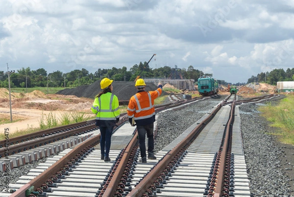 Fototapeta Rear view, two Asian railway engineers or workers wearing reflective vests inspect railroad track construction at station, outdoors.