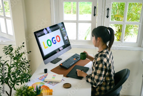 Fototapeta Graphic designer creating logo on digital tablet in bright modern workspace with computer and color palette