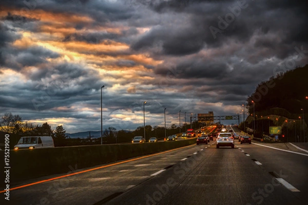 Obraz A dramatic sky with dark, fiery clouds hangs over a busy highway during the evening commute. The image captures the movement of vehicles on the interstate with bright orange and white lights.