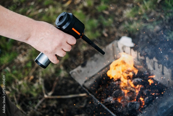 Fototapeta Close-up of a hand using a battery-powered blower to stoke flames in a metal firepit. Fire starter technology, portable blowers, camping gadgets, outdoor innovation