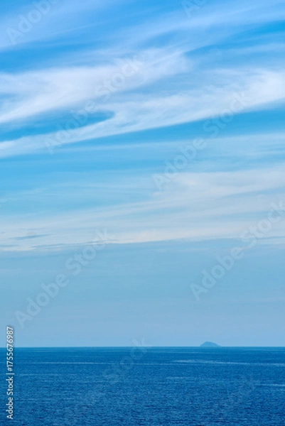 Fototapeta Deep blue ocean under an expansive sky with wispy clouds. A lonely island on the horizon adds a sense of scale, solitude, and distant destination.