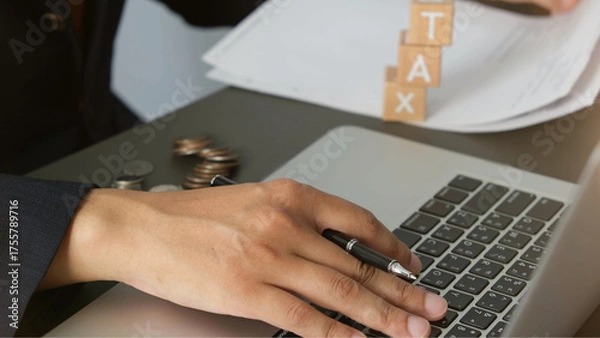Fototapeta Businessman working on tax documents with laptop, coins, and wooden blocks spelling TAX, showing financial planning and focus