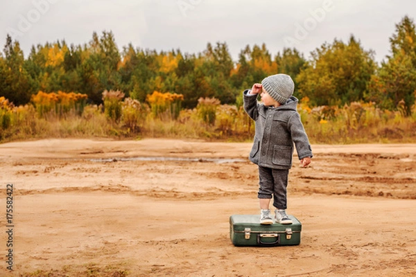 Fototapeta a boy in panties and a gray coat is standing on an old suitcase in the middle of a sandy country road, raising his hand to his forehead