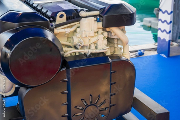 Fototapeta Detailed view of a large marine engine displayed on a black metal frame indoors, featuring black protective covers and white pipes, set against a blue floor background.