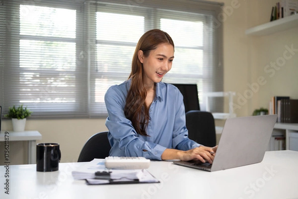 Fototapeta Smiling businesswoman working on laptop at modern office desk with natural daylight.