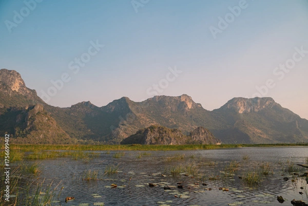 Fototapeta The mountain looms over a green landscape where a swamp meets a tranquil lake, water reflecting the sky, a single cloud drifting in peaceful, untouched nature.