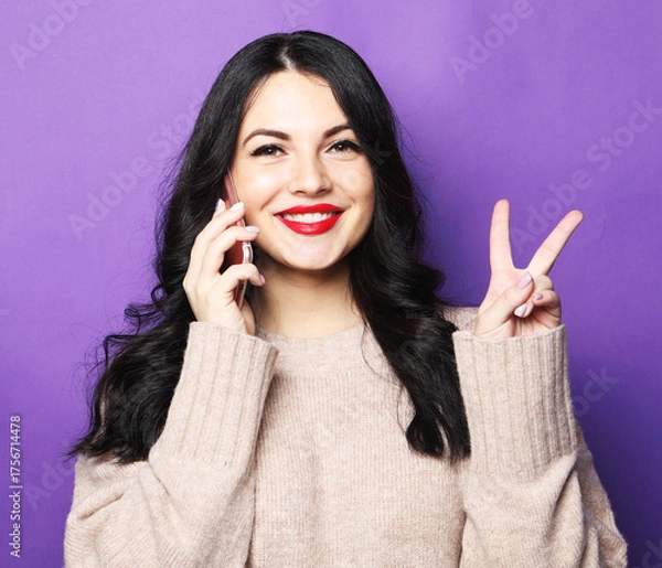Obraz Beautiful smiling brunette woman talking on mobile phone and showing v-sign over purple background.
