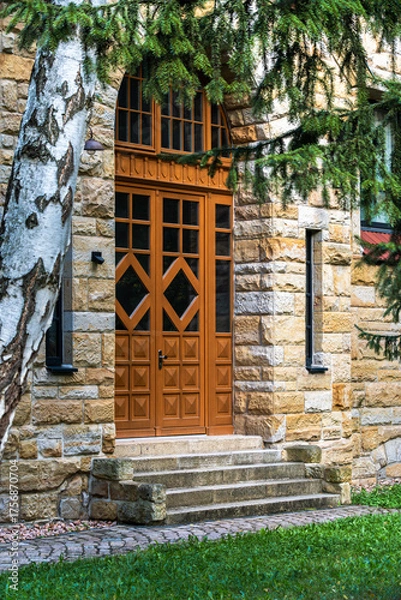 Fototapeta Massive stone facade with an ornate wooden door and steps, framed by greenery and a birch trunk, photographed in daylight in Gotha