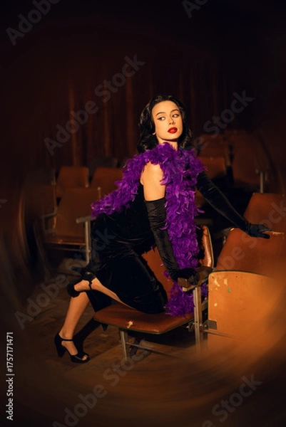 Fototapeta Elegant woman in black dress with purple feather boa sits in empty theater during evening event