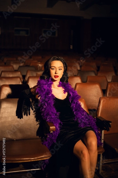 Fototapeta Elegant woman in black dress with purple feather boa sits in empty theater during evening event
