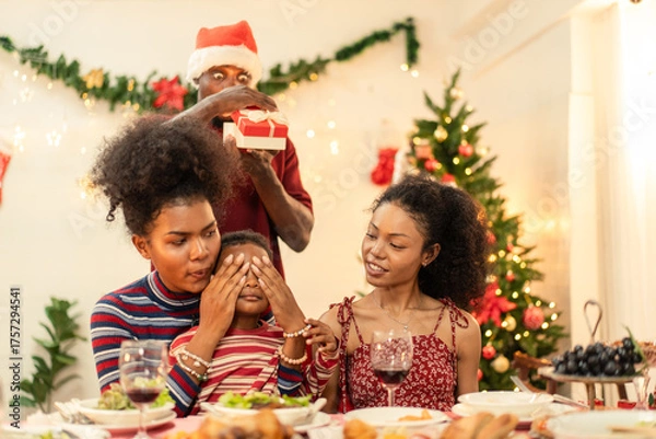 Obraz Happy Black family celebrating Christmas dinner as a child's eyes are covered for a surprise gift. Father prepares the present. Festive surprise, holiday joy, and family tradition during the winter