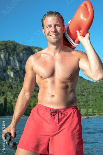 Obraz young, sexy, muscular man is doing lifeguard - he is watching at the bay for the safety. he wears red swimming trunks and is holding an red rescue buoy and a binoculars. 