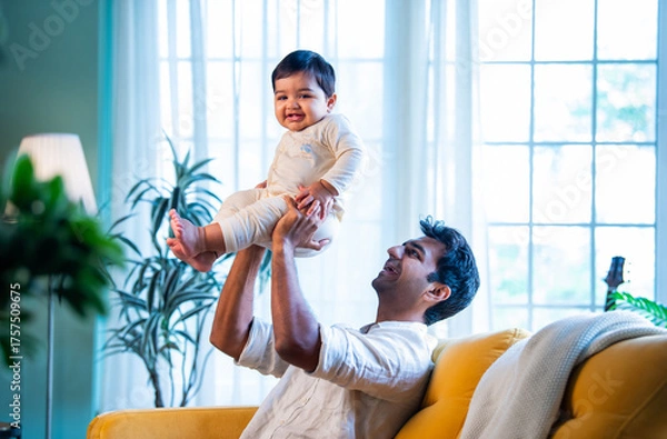 Fototapeta Indian Playful father and baby boy smiling while playing on living room sofa at home