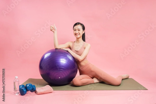 Obraz Smiling asian woman with a thumbs up resting on a purple exercise ball after her workout