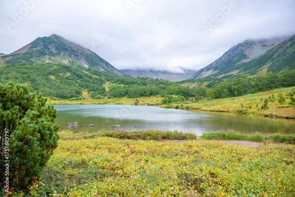Fototapeta Scenic landscape of lush green valley with clear lake and majestic mountain peaks beneath cloudy sky capturing serene natural beauty and tranquil atmosphere in summer.