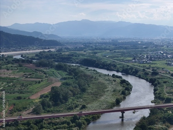 Obraz A river runs through a valley with a bridge over it. The bridge is red and spans the width of the river. The valley is surrounded by mountains, and the sky is clear and blue