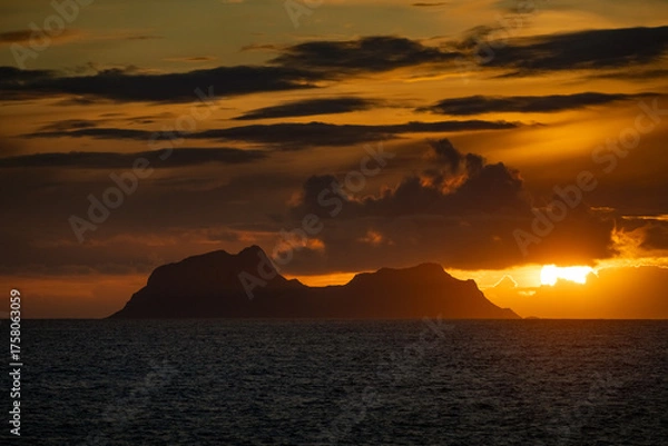 Fototapeta Silhouette of Mosken island (Moskenesøy) and its dramatic mountain peaks at sunset, with a fiery orange sky and dark, moody clouds casting a glow over the Norwegian Sea
