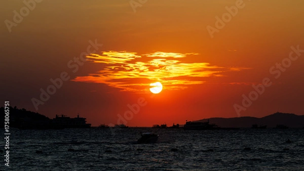 Fototapeta round sun sets over the sea, painting the sky in intense orange and deep red hues. Several boats are anchored and sailing across the water below, forming sharp, contrasting silhouettes.