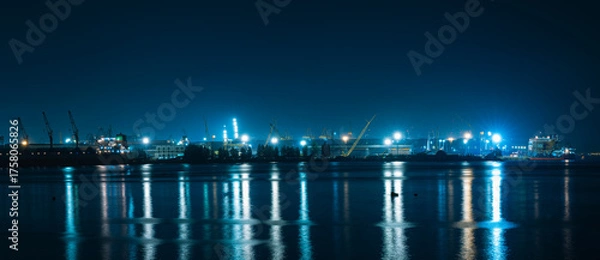 Fototapeta panoramic image of an industrial port at night. Bright blue-white lights from cranes and structures reflect in long, straight streaks on the deep blue water, 