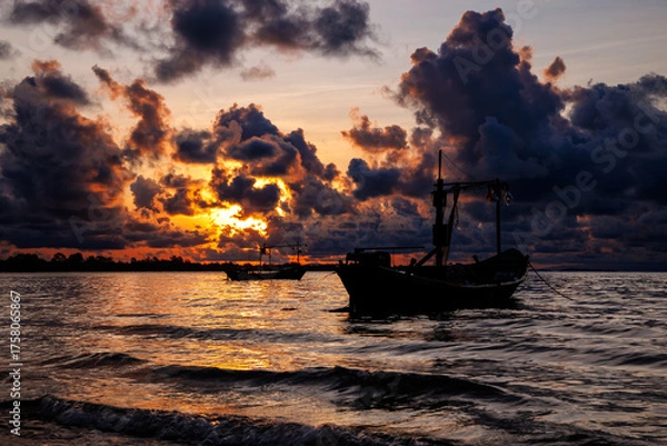 Fototapeta A silhouette of two fishing boats is set against the shimmering water as the sun dips behind massive, dark, and intense orange clouds.