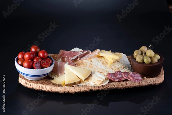 Obraz pane carasau con salame, formaggio e olive su vassoio in sughero sfondo nero