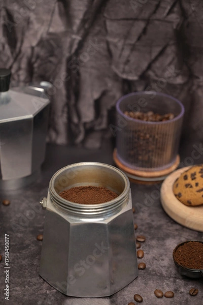 Obraz Geyser coffee maker with ground coffee standing on the table. Coffee beans in a glass. Coffee in a black spoon. Oatmeal cookies with chocolate chips on a wooden board. Dark background. Still life