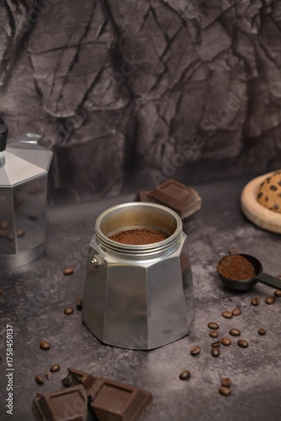 Obraz Geyser coffee maker with ground coffee standing on the table. Coffee in a black spoon. Pieces of broken dark chocolate. Spilled coffee beans. Oatmeal cookies on a wooden board. Dark background