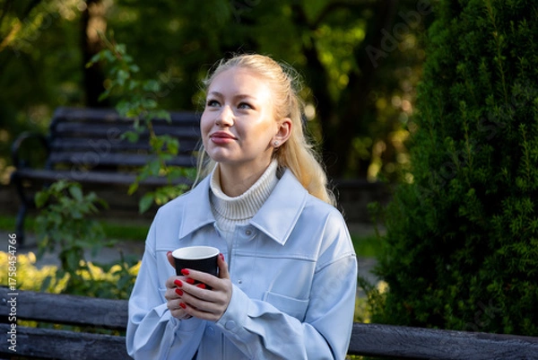 Obraz Cheerful young blonde woman in light blue jacket enjoys a quiet moment with a hot drink, smiling and looking up while sitting on a bench surrounded by green trees and sunlight