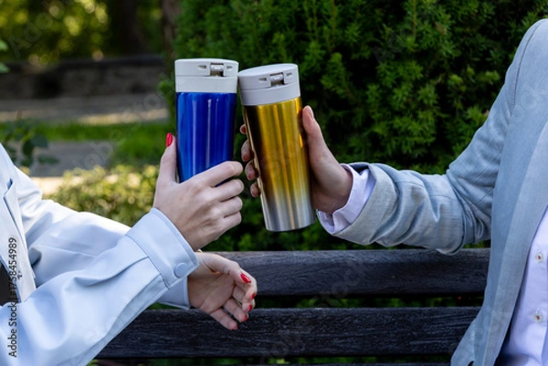 Obraz Close-up of two people in light jackets clinking reusable thermos mugs one blue, one gold while sitting on a wooden bench in a green city park on a sunny autumn day