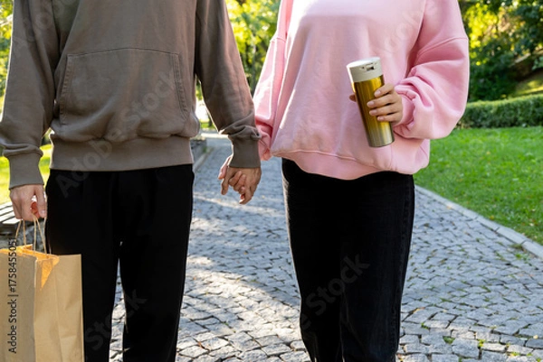 Obraz Close up of a couple walking together on a cobblestone path in a park, holding hands. One holds a paper shopping bag, the other carries a gold thermos, both dressed in cozy hoodies