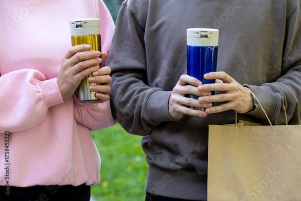 Obraz Close-up of two people in casual pink and brown hoodies holding colorful reusable travel mugs gold and blue while standing in a park, with a paper shopping bag in hand
