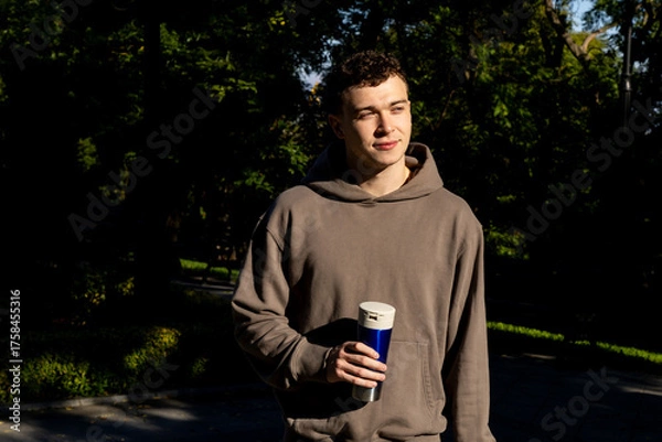 Obraz Young man in a taupe hoodie holds a reusable blue coffee tumbler while standing in a sunlit green park, gazing peacefully into the distance on a calm autumn day