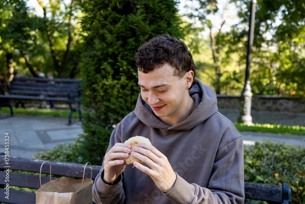 Obraz Young man in a brown hoodie sits on a wooden bench in a sunny park, holding a sandwich with both hands and smiling as he gets ready to eat, surrounded by greenery and a paper bag
