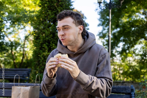 Obraz Young man in a brown hoodie sits on a wooden bench chewing a sandwich, smiling with full cheeks, surrounded by greenery and soft sunlight on a relaxed autumn day