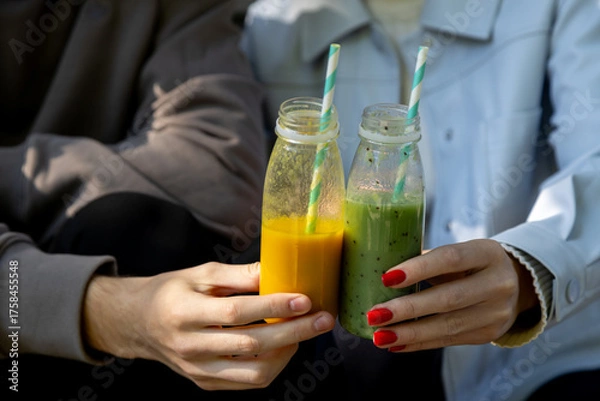 Obraz Hands of a man and woman clink together two eco-friendly glass bottles filled with fresh smoothies one vibrant orange and the other green with kiwi seeds each with a striped paper straw, against a sof
