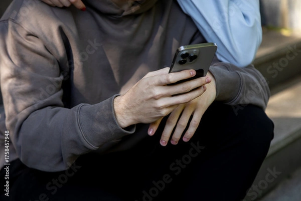 Obraz Detail shot of a mans hands holding a modern smartphone as he sits on stone steps, with a womans arm affectionately resting on his shoulder, suggesting a shared digital experience