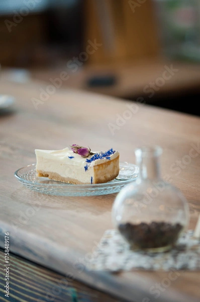 Fototapeta Close-up of a delicious cheesecake slice served on a glass plate, decorated with edible flowers — rosebuds and blue cornflower petals. Elegant dessert presentation on a wooden table, perfect for illus