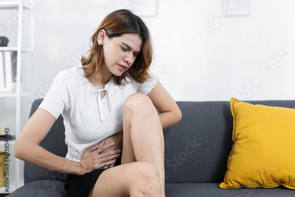 Fototapeta women sitting on sofa with hand on stomach, expressing abdominal pain or cramps, symbolizing digestive issues, period pain, bloating, food intolerance and need for health care.