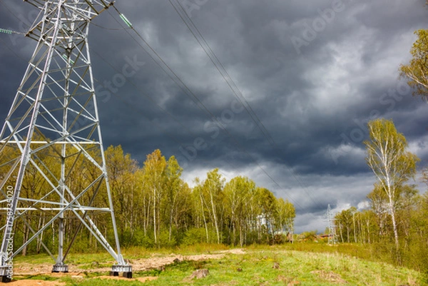 Fototapeta Sturdy electricity pylon commands the foreground, silhouetted against a brooding, stormy sky. Vibrant green spring foliage lines the horizon