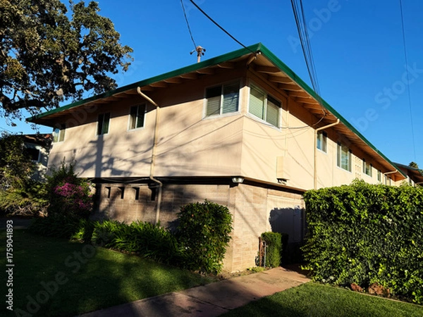 Fototapeta Two-story low-rise beige apartment building with green roof, multiple windows, and landscaped garden with hedges and flowering plants under clear blue sky in a suburban residential area