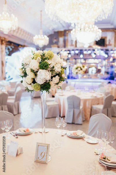 Fototapeta Tall vase with white and green flowers stands on round dinner table number nine