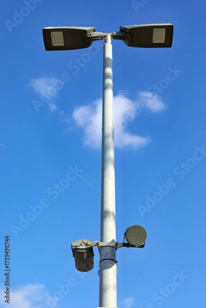 Fototapeta A tall street light pole with two large modern luminaires against a bright blue sky. Lower on the pole, two additional devices are mounted, possibly sensors or cameras.
