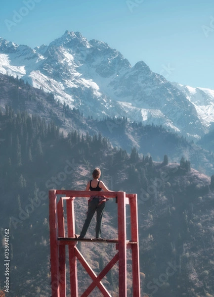 Fototapeta a woman standing on a wooden observation deck, admiring the stunning view of snow capped mountains and forest on a sunny day.