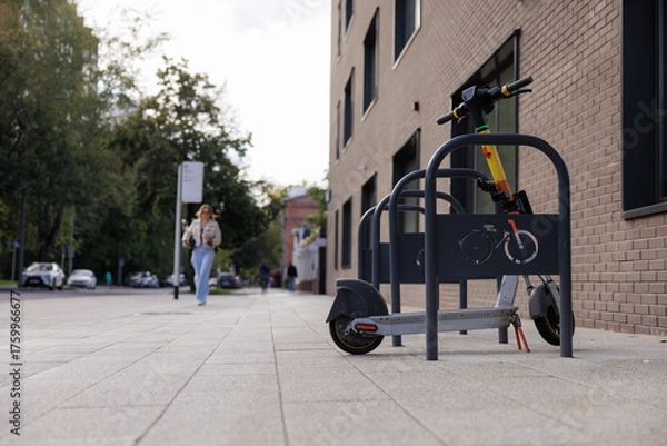 Fototapeta Scooter is secured at a bike rack on a city sidewalk while a woman walks by in casual attire on a sunny afternoon.