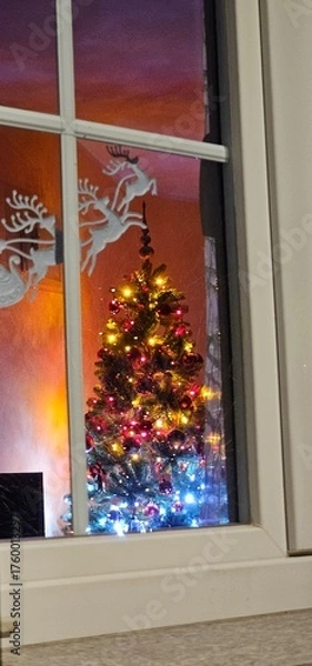 Fototapeta A lit Christmas tree with Christmas decorations through a window inside a house immersed in the Christmas holidays