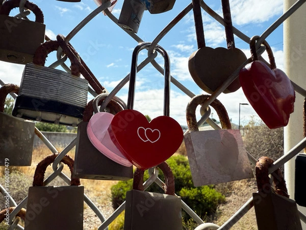 Fototapeta Multiple padlocks including red heart lock hanging on wire fence with natural background and clouds, expressing concept of love, memory, promise, and human emotion