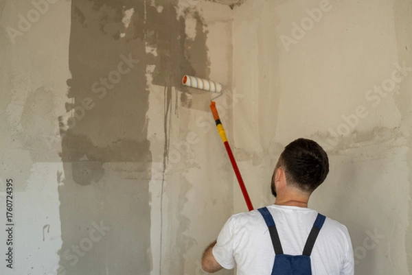 Obraz A bearded man in a work uniform is priming the walls after applying gypsum plaster.