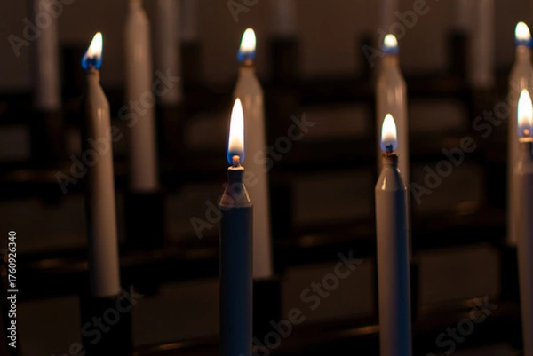 Fototapeta Low-light photo of several tall, white votive candles burning brightly in a church or sanctuary, creating a solemn and peaceful atmosphere