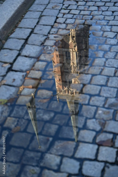 Fototapeta Reflection of Gothic cathedral towers in a rain puddle on cobblestone pavement. The image highlights timeless architecture, European charm. Ideal for travel magazines, design projects, posters.
