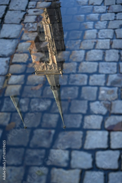 Fototapeta Reflection of Gothic cathedral towers in a rain puddle on cobblestone pavement. The image highlights timeless architecture, European charm. Ideal for travel magazines, design projects, posters.